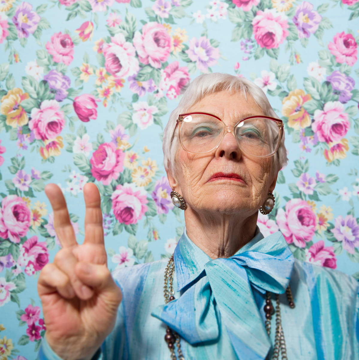 Äldre dam i kort hår och turkos blus håller upp peacetecken med fingrarna mot en turkos bakgrund med rosa blommor
